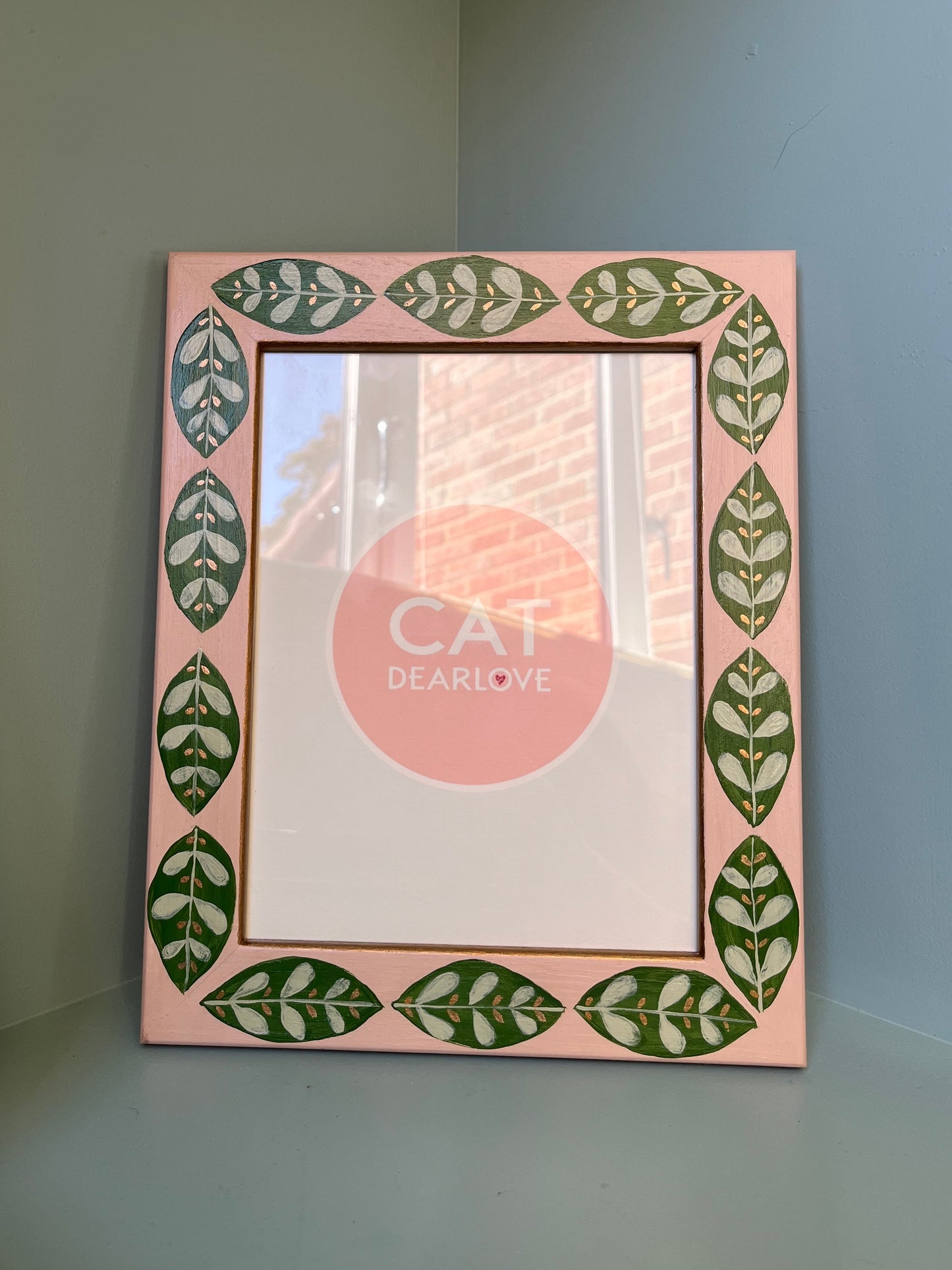 Nude pink hand painted frame with green leaf motif and gold finish. Frame is sitting on a shelf. 